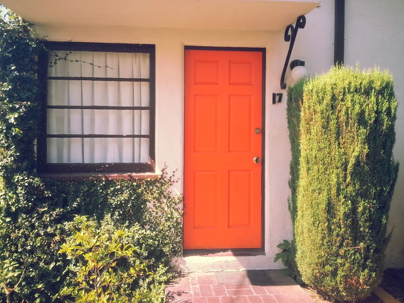 A house with a bright orange door, a window with a grid pattern, and a tall green hedge.