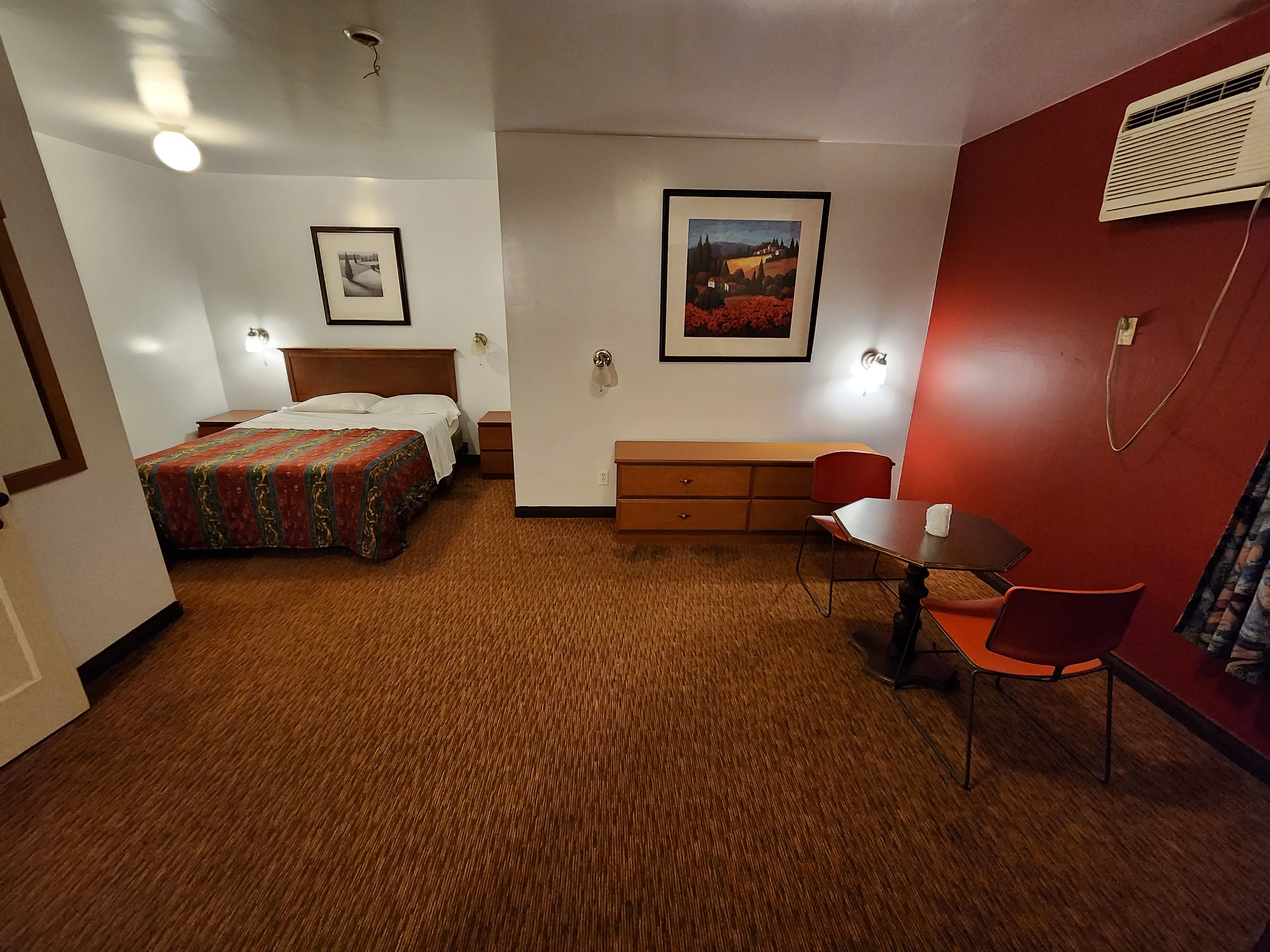 A hotel room with a bed, a desk, a chair, a dresser, and two framed pictures on the walls.