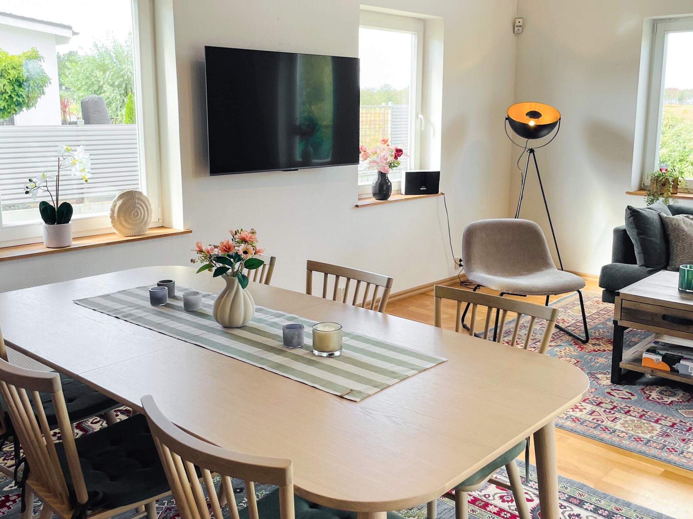 Spiseplads med bord, stole, en vase med blomster, et fladskærms-tv ophængt på væggen og en gulvlampe.