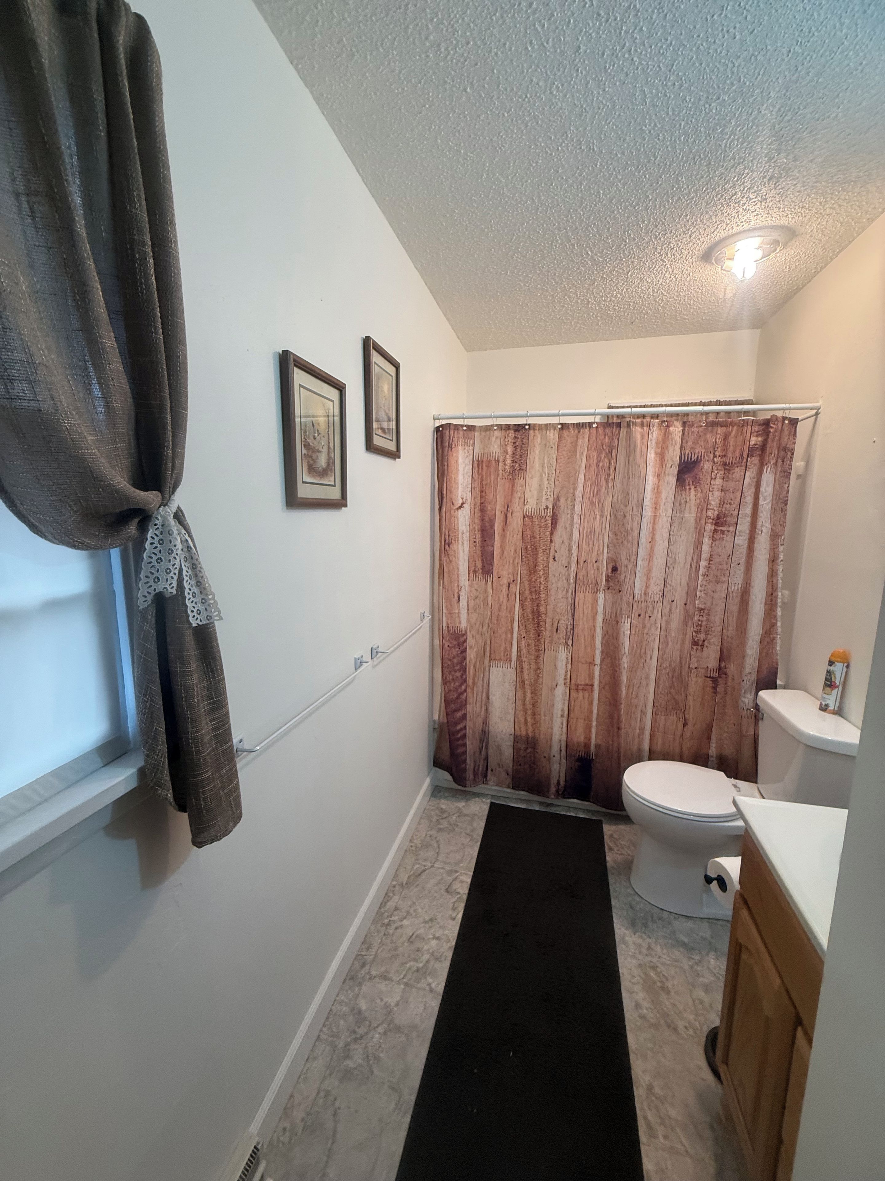 A bathroom with a wooden shower curtain, a toilet, a sink, and a window with a towel hanging.