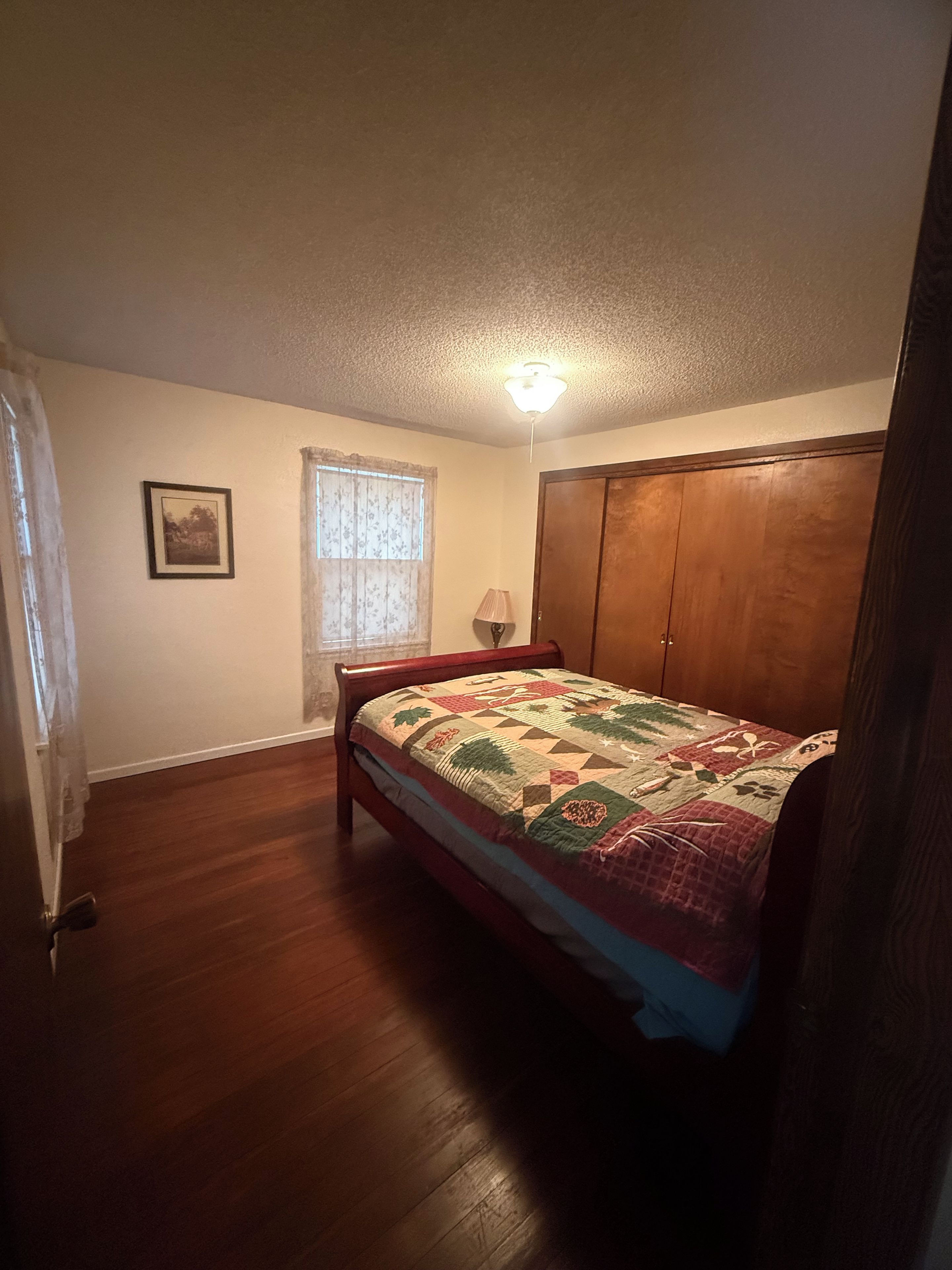 A bedroom with a wooden bed, a window with curtains, a ceiling light, and a door.