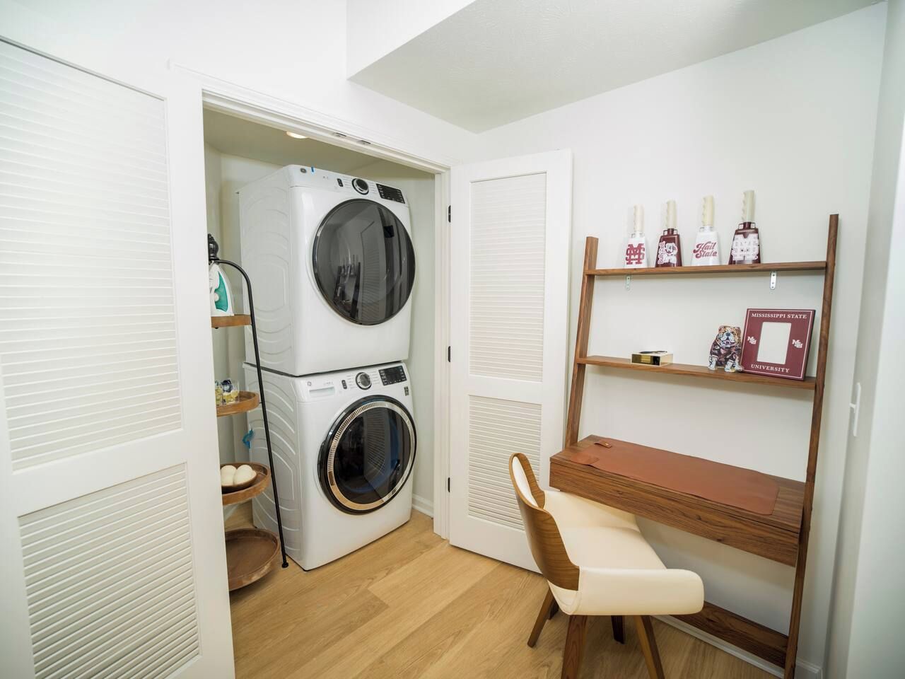 A laundry room with stacked washers and dryers, a wooden chair, and a shelf with bottles and a framed item.