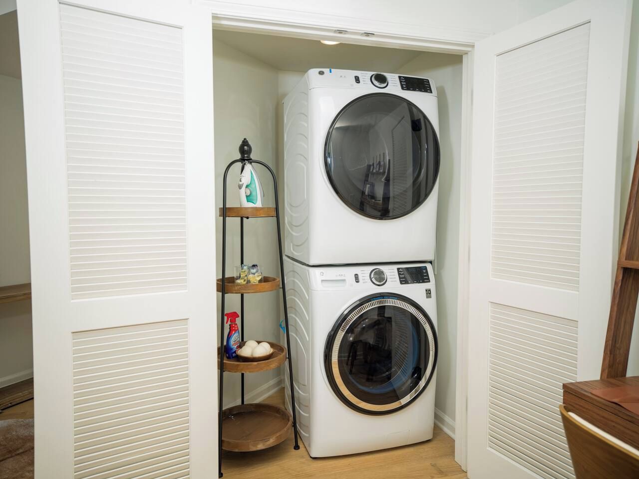 A compact laundry room with a stacked washer and dryer, a shelf with cleaning supplies, and a wooden floor.