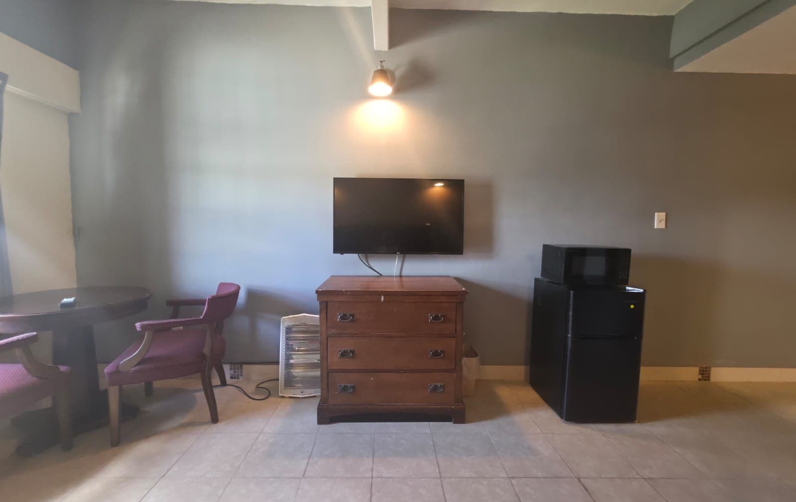 A room with a flat-screen TV mounted on the wall, a wooden dresser, a refrigerator, and a dining table with chairs.