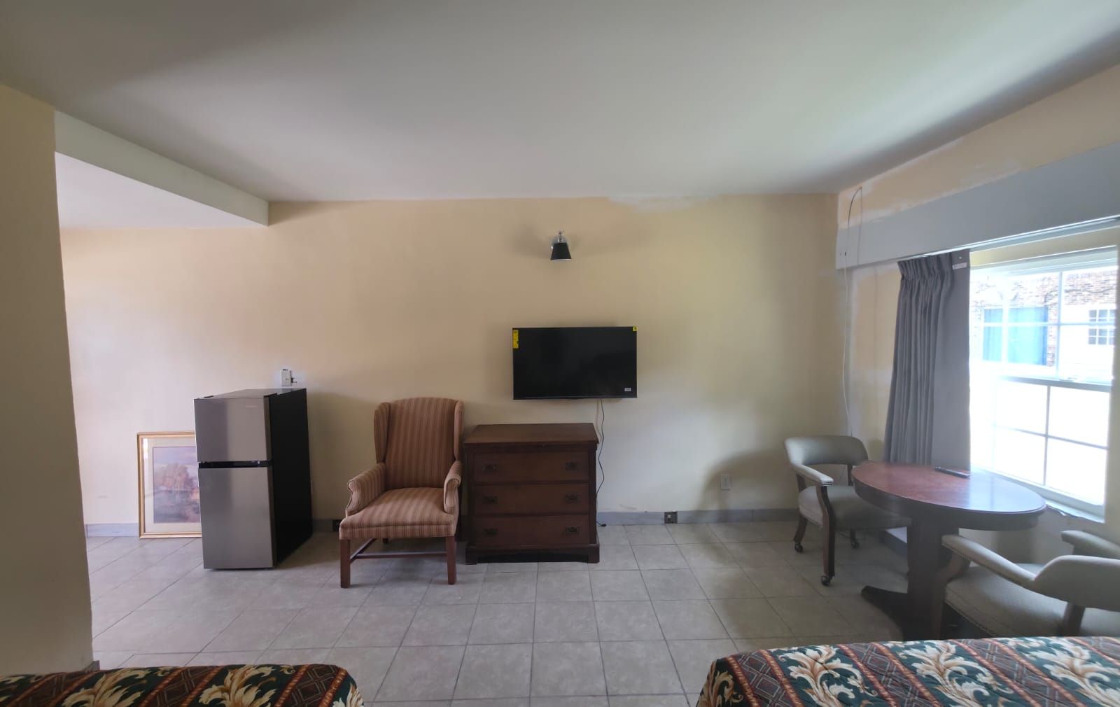Una habitación con una televisión de pantalla plana montada en la pared, una mesita con sillas, un sofá y un refrigerador.