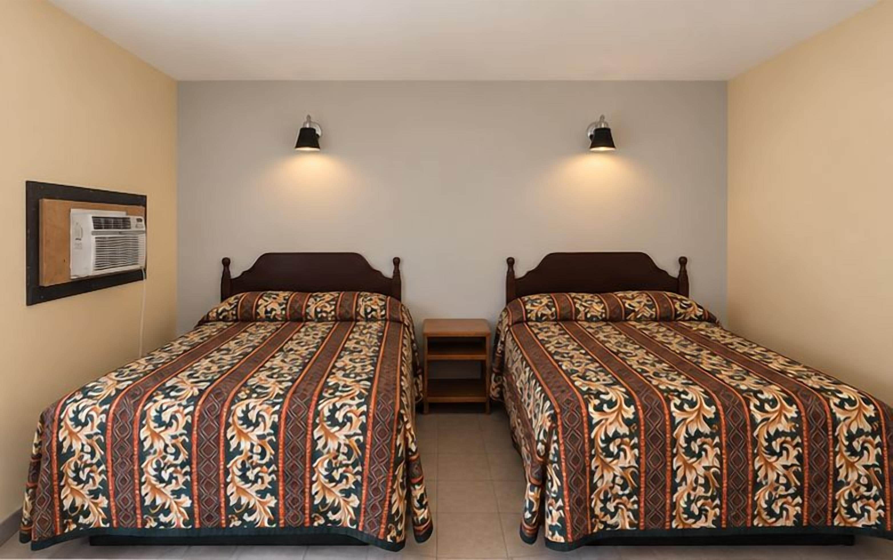 A room with two single beds, each with a patterned bedspread, a small wooden nightstand, and a wall-mounted air conditioning unit.