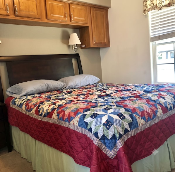 A bedroom with a bed, a nightstand, wooden cabinets, a wall-mounted lamp, and a window with blinds.