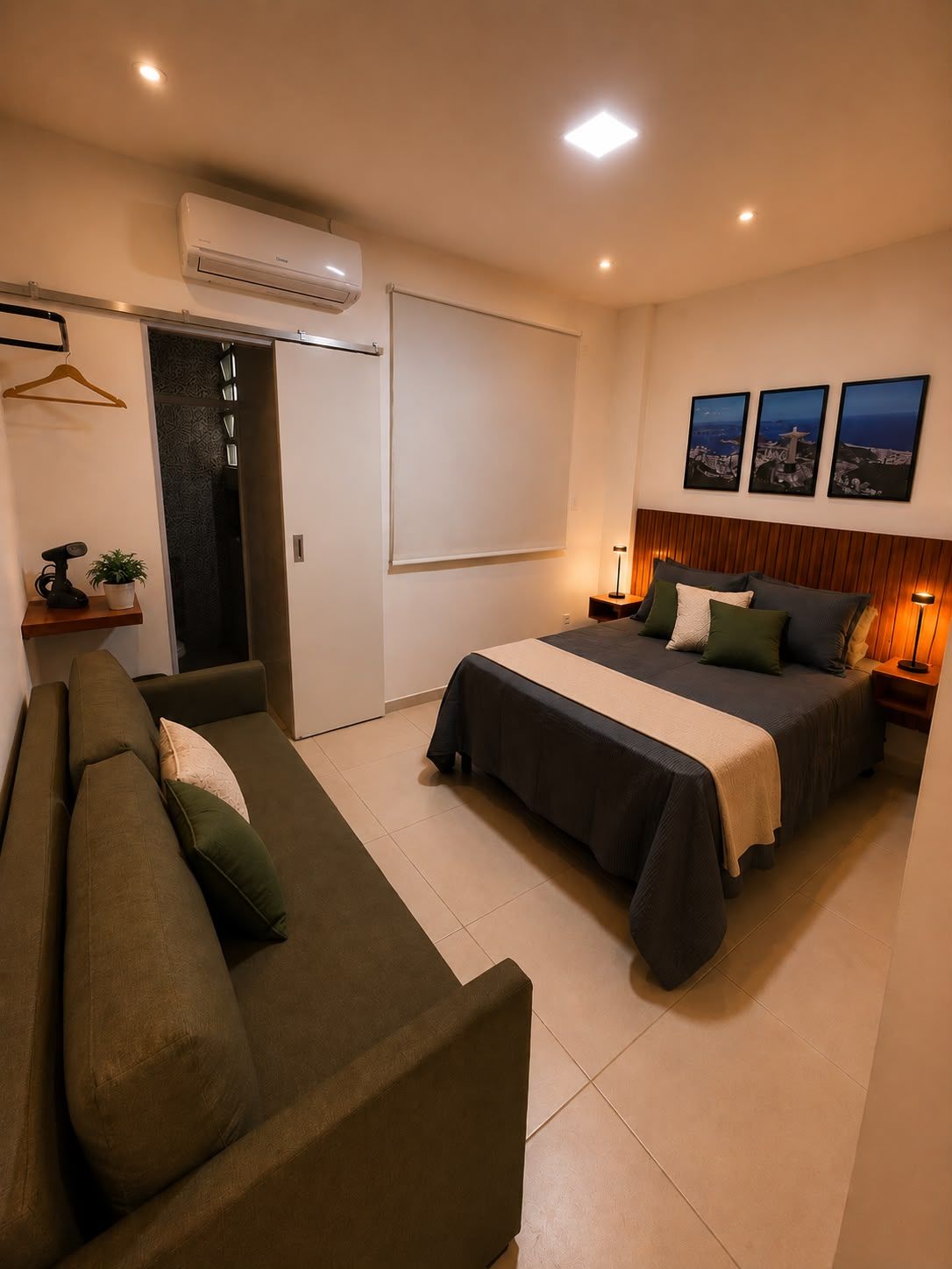 A modern bedroom with a bed, a sofa, an air conditioning unit, and a door leading to a bathroom.