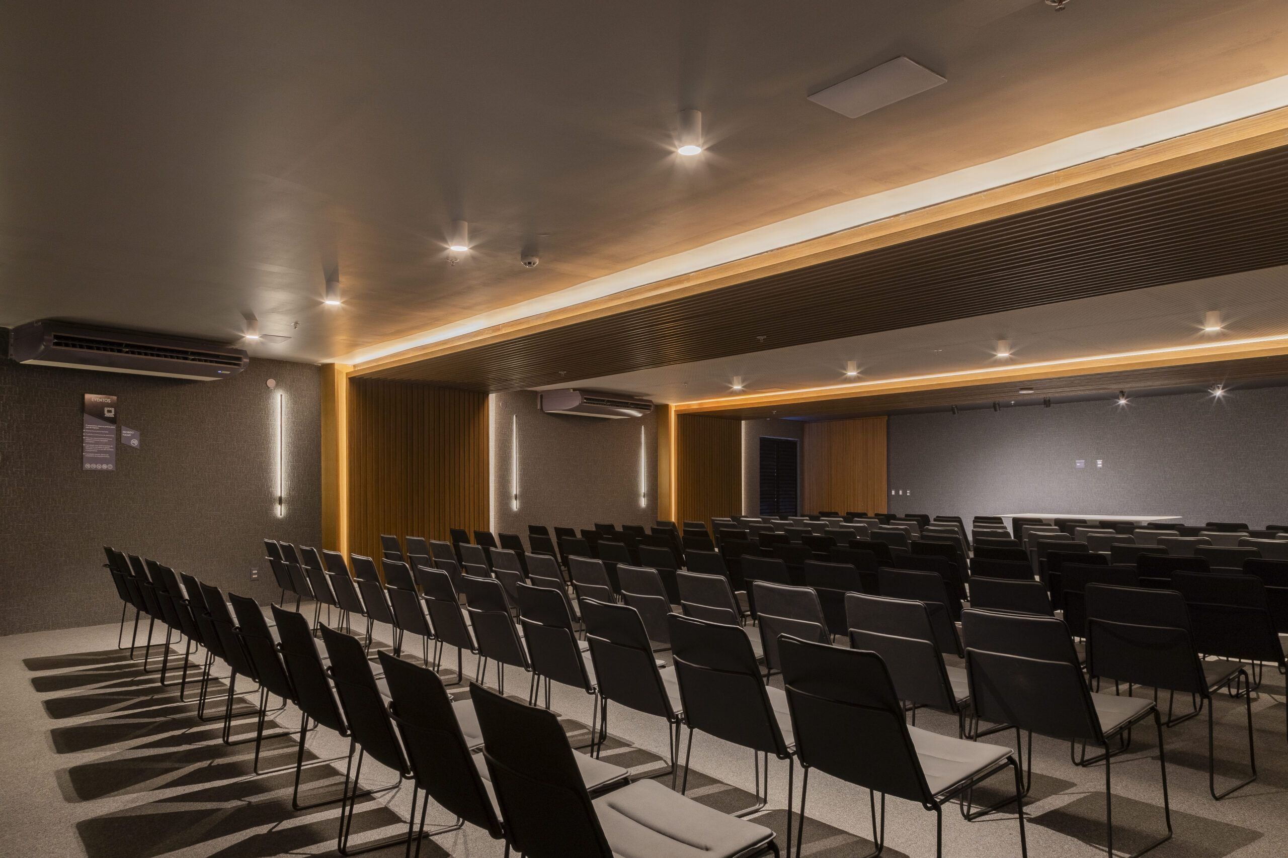 Un salón de conferencias con hileras de sillas, un revestimiento de pared de madera y luces en el techo.