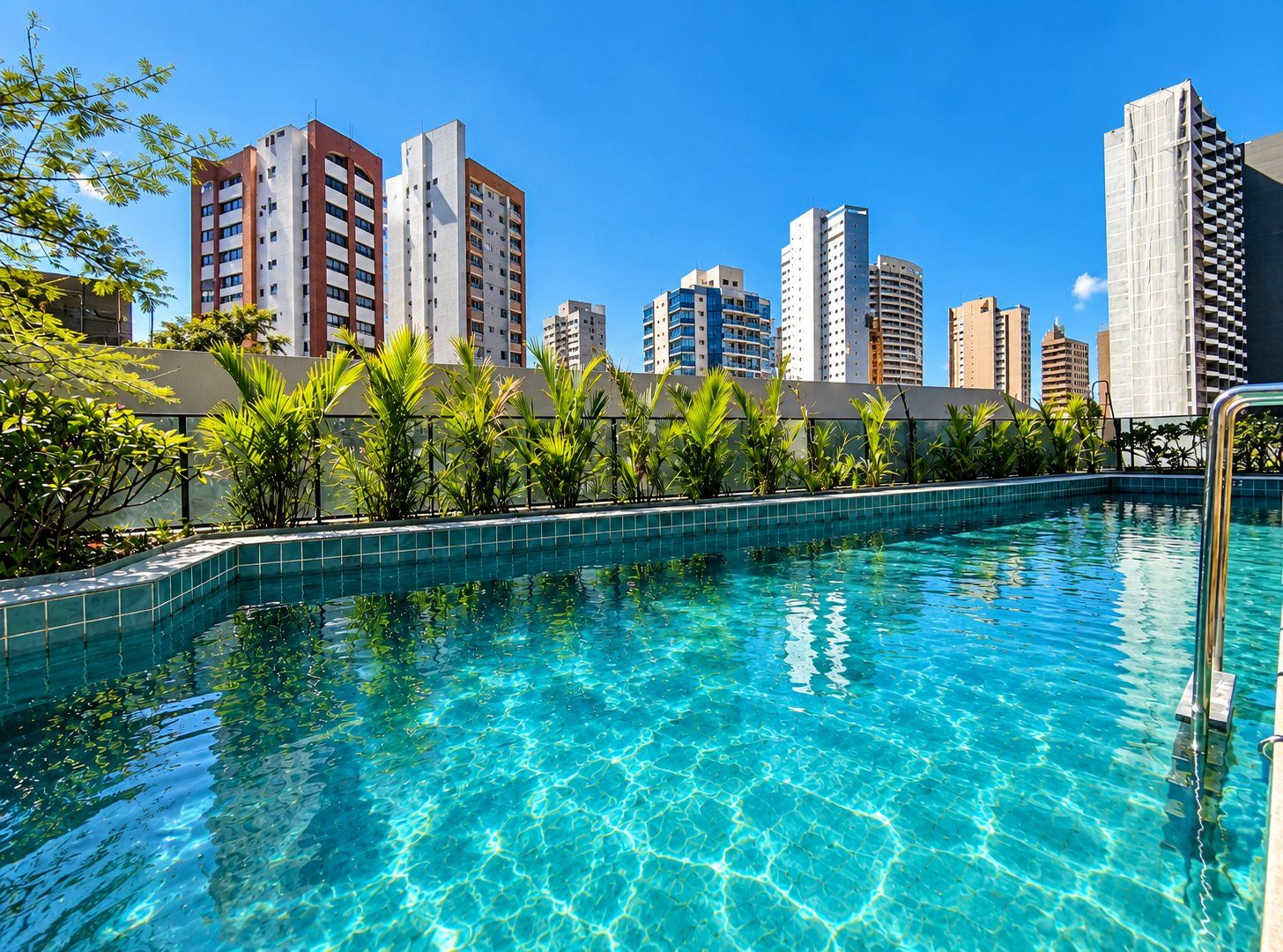 Una piscina con agua azul cristalina, rodeada de follaje y altos edificios al fondo.