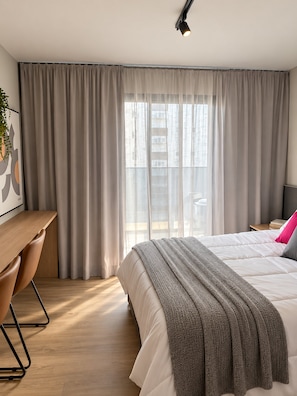 Habitación de hotel con cama, escritorio y un amplio ventanal con cortinas.