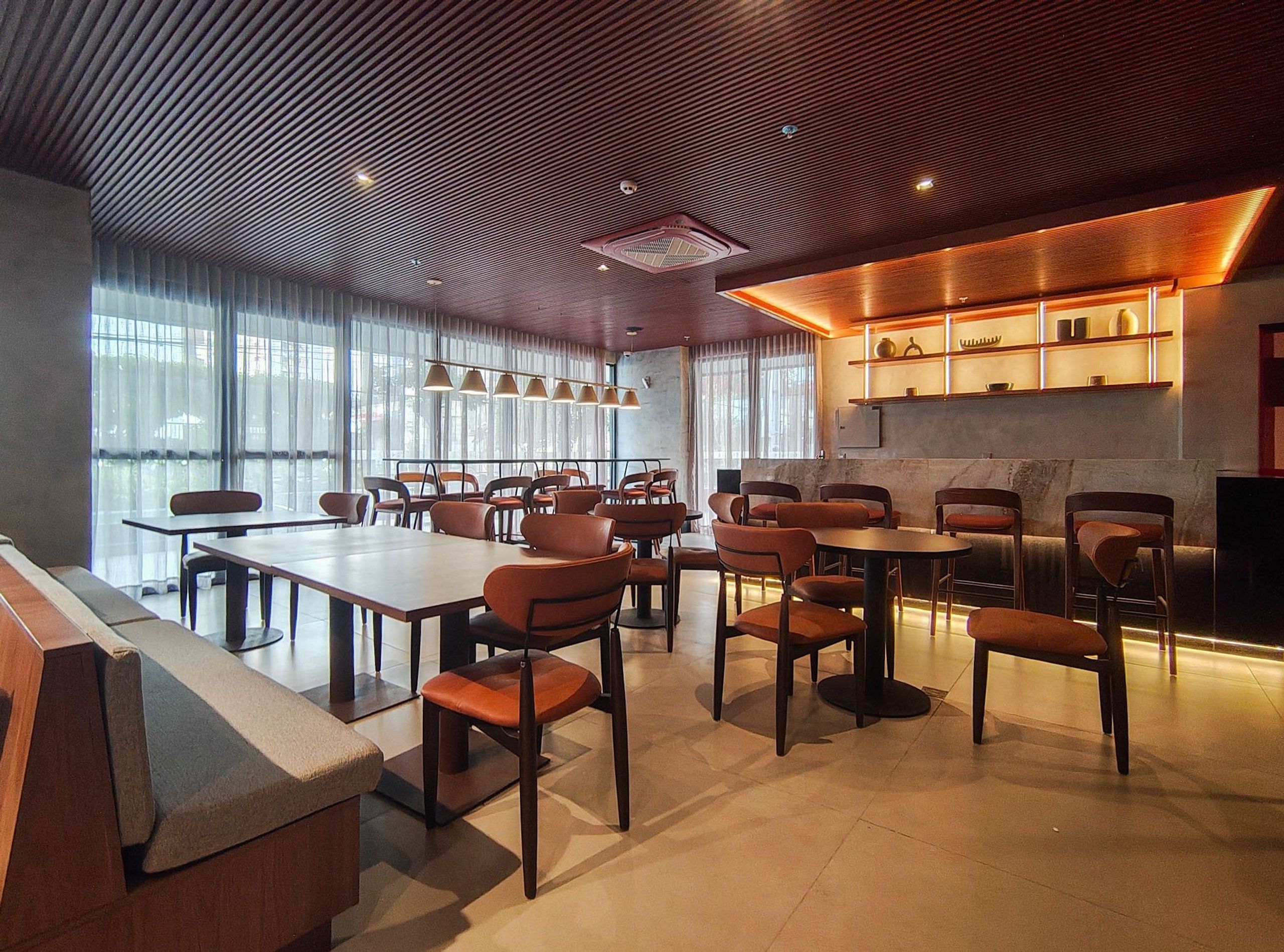 A modern dining area with multiple tables and chairs, large windows, and a kitchen area with shelves.