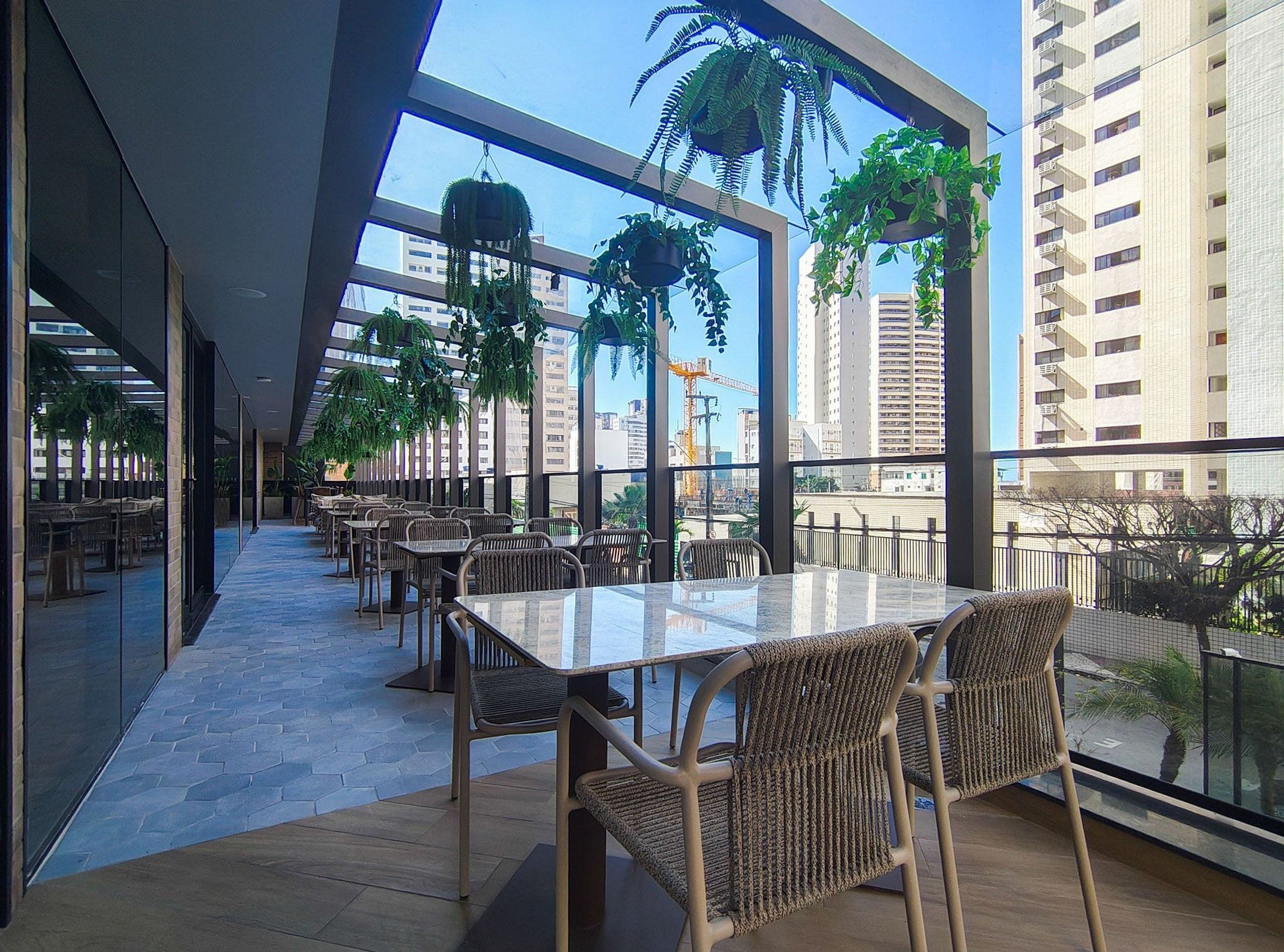 Un comedor interior con mesas y sillas, ventanas amplias y plantas colgantes.