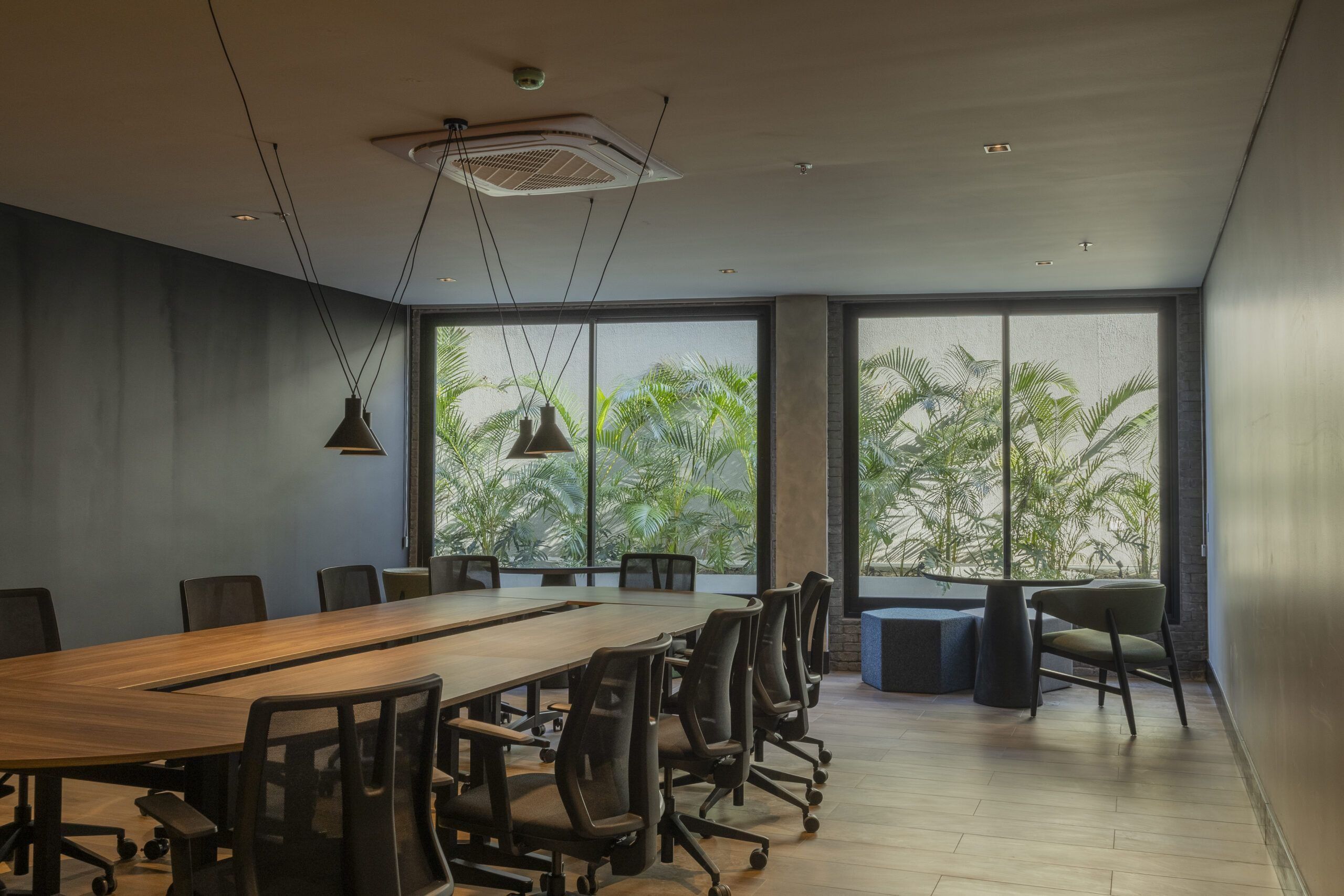 A modern conference room with a long table, chairs, and large windows with a view of greenery.