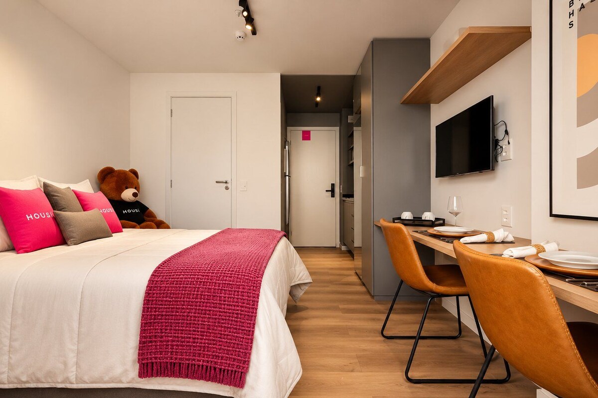 Habitación de hotel con cama, escritorio con sillas, televisor y un oso de peluche.