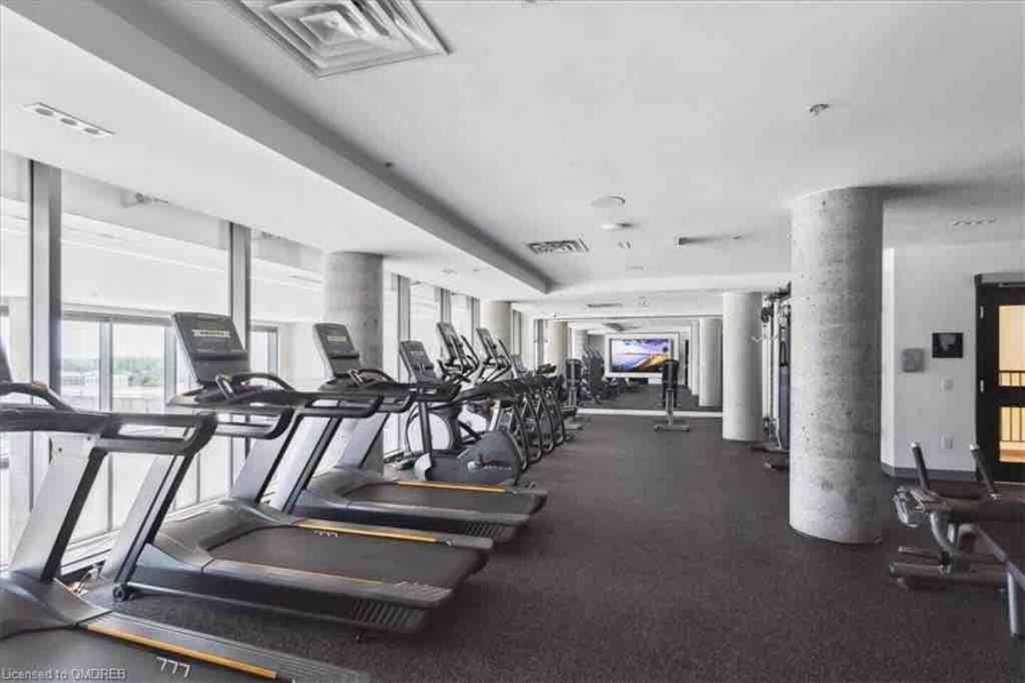 A gym with treadmills and exercise equipment, large windows, and a television screen.