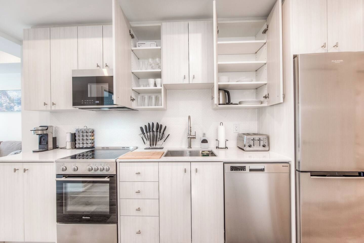 A modern kitchen with white cabinetry, a stainless steel refrigerator, a built-in oven, a dishwasher, and a coffee machine.