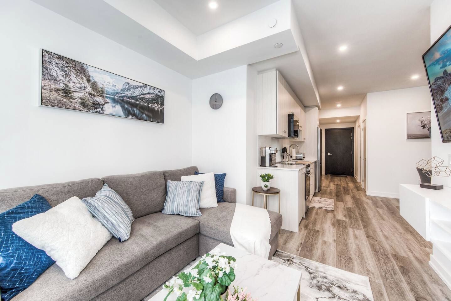 A modern living room with a grey sofa, a coffee table with flowers, a kitchen area with white cabinets, and a dining area with a wooden floor.