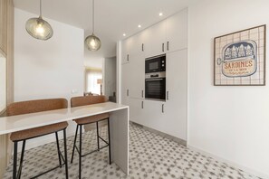 Eine moderne Küche mit weißer Arbeitsplatte, zwei Barhocker, einer Pendelleuchte und einem eingebauten Backofen.