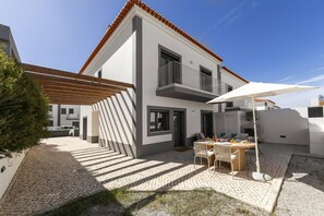 Ein modernes, zweigeschossiges Haus mit einer Terrassenfläche, Außenbestuhlung und einem Auto auf der Auffahrt.