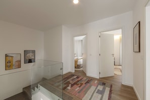 A modern interior with a glass railing staircase, a patterned rug, and a hallway leading to a bedroom with a bed and a nightstand. - Beachside Hideaway - Elegant Coastal Getaway (Peniche)