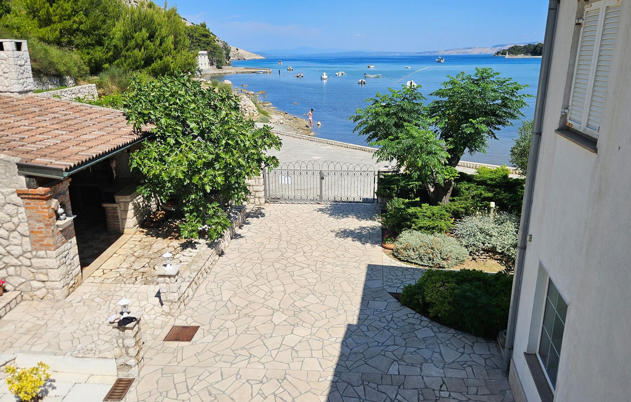 A stone-paved courtyard with a view of the sea, a gate, and surrounding greenery.