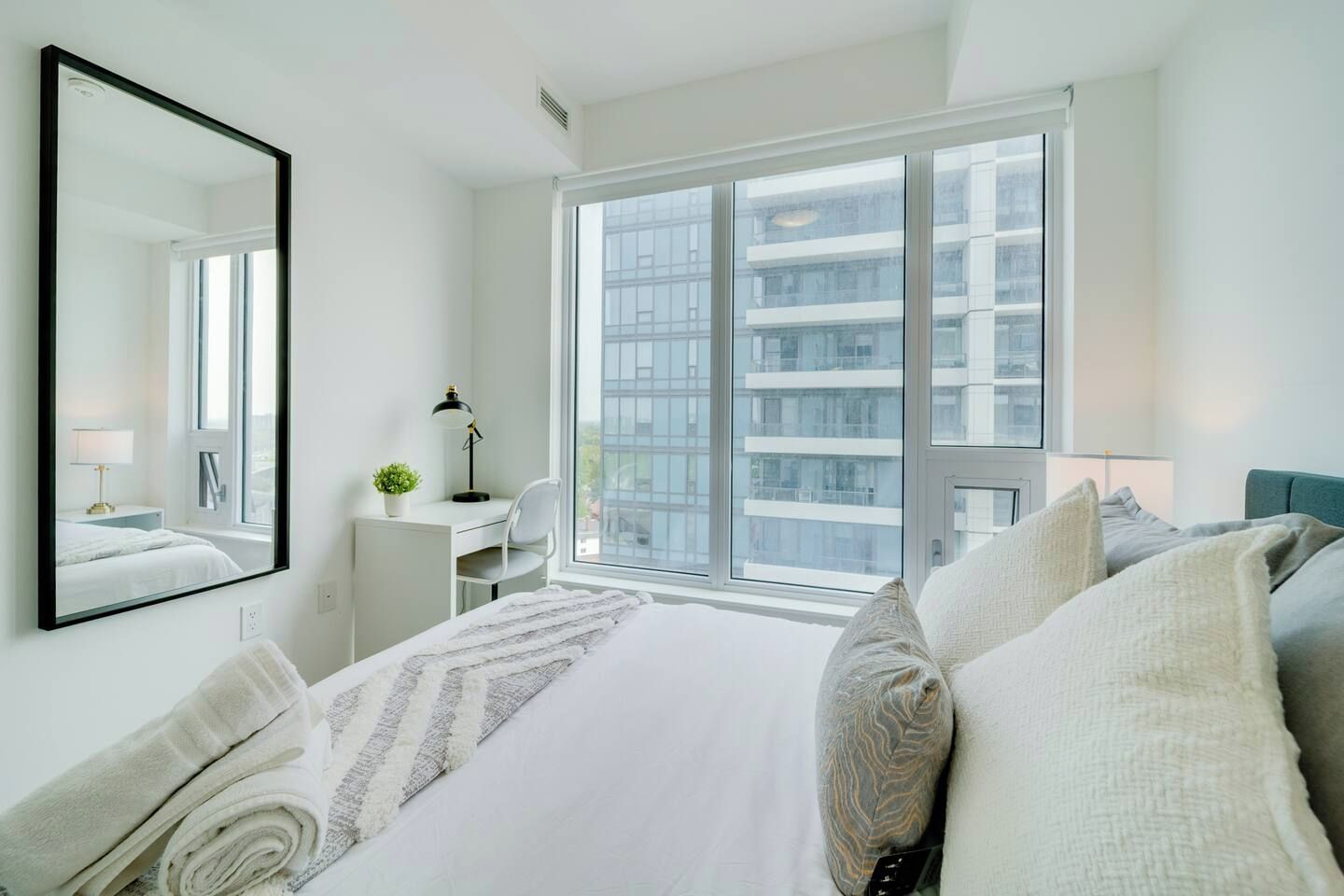 A modern hotel room with a large bed, a desk, a chair, and a large window offering a view of the city.