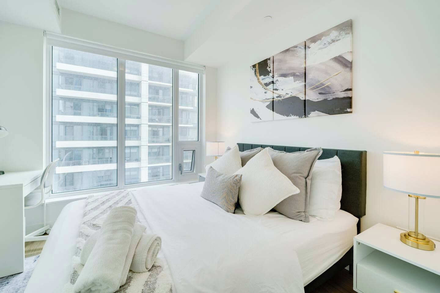 Habitación de hotel con una cama grande, un escritorio, una silla, un ventanal con vistas a edificios y un cuadro en la pared.