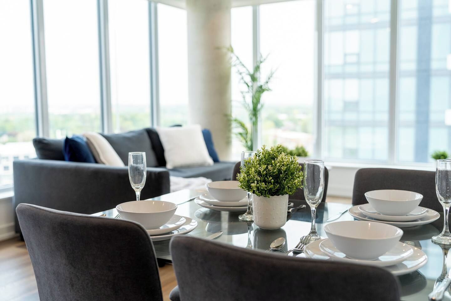 A dining area with a table set for four, including plates, bowls, cutlery, and glasses. There is a potted plant on the table, and the room has large windows with a view of the city.