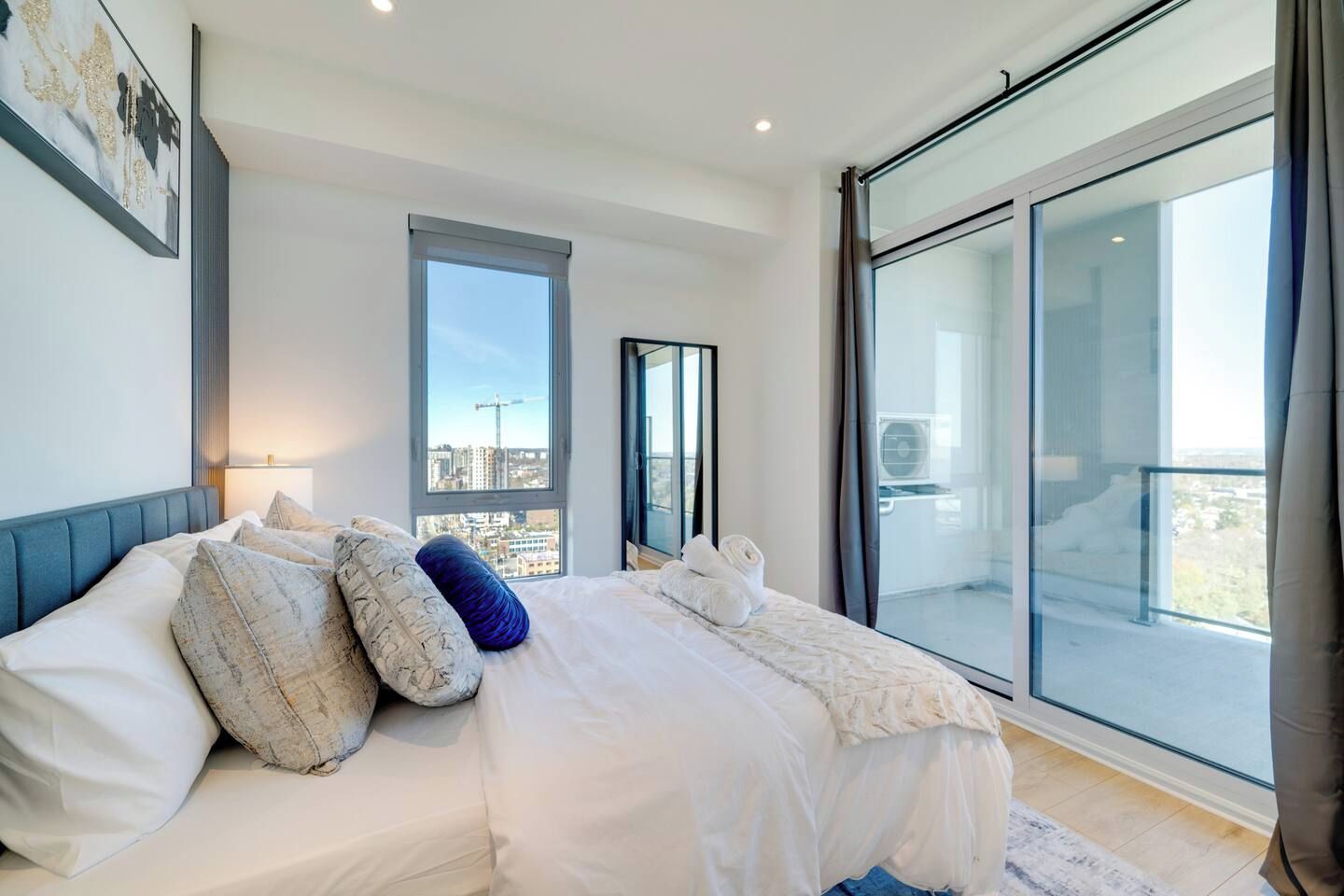 Un dormitorio moderno con una cama grande, vistas de la ciudad a través de una ventana y un balcón con puerta de cristal.
