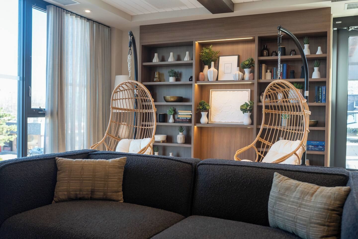 A modern living room with a dark grey sofa, two wicker chairs, a wooden bookshelf, and a large window with curtains.