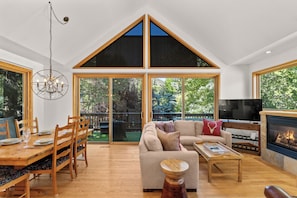 Amplia sala de estar con techos altos, chimenea, comedor con mesa y sillas de madera, y ventanas grandes con vistas al exterior.