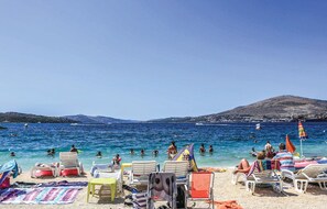 Ein Strand mit Liegestühlen und Sonnenschirmen, blaues, klares Meer und eine hügelige Landschaft im Hintergrund.