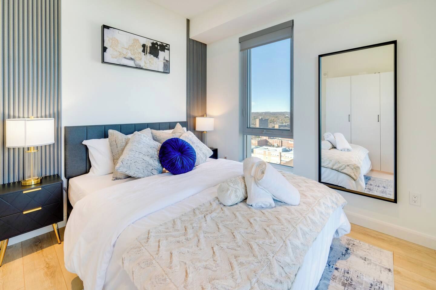 A modern bedroom with a large bed, a nightstand, a bedside lamp, a window with a city view, and a mirror.