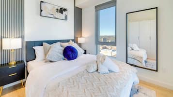 A modern bedroom with a large bed, a nightstand, a bedside lamp, a window with a city view, and a mirror.