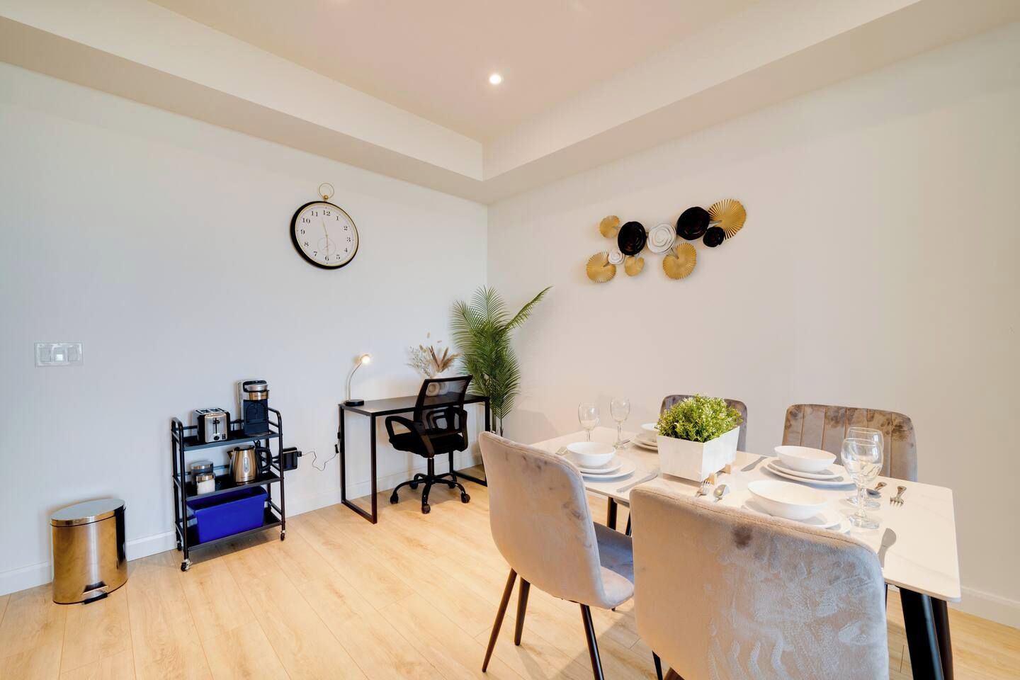 A modern dining area with a wooden floor, a dining table set for two, a black office chair, a small desk with a lamp, and a wall clock.