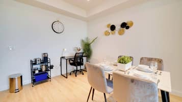 A modern dining area with a wooden floor, a dining table set for two, a black office chair, a small desk with a lamp, and a wall clock.