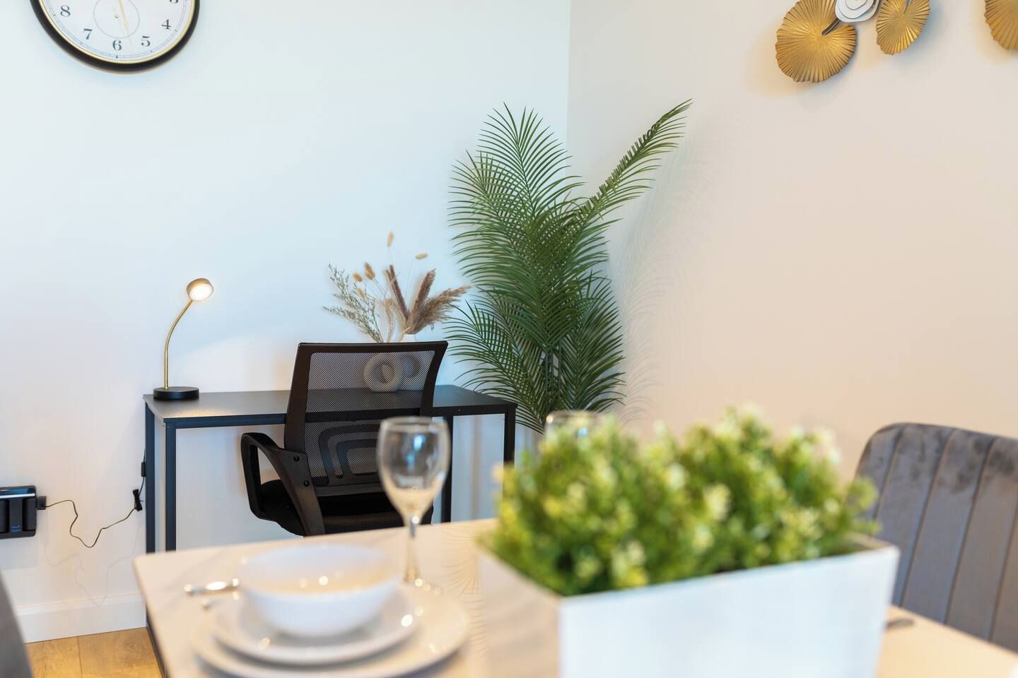 A dining area with a table set for two, a laptop on a desk, a potted plant, and a wall clock.