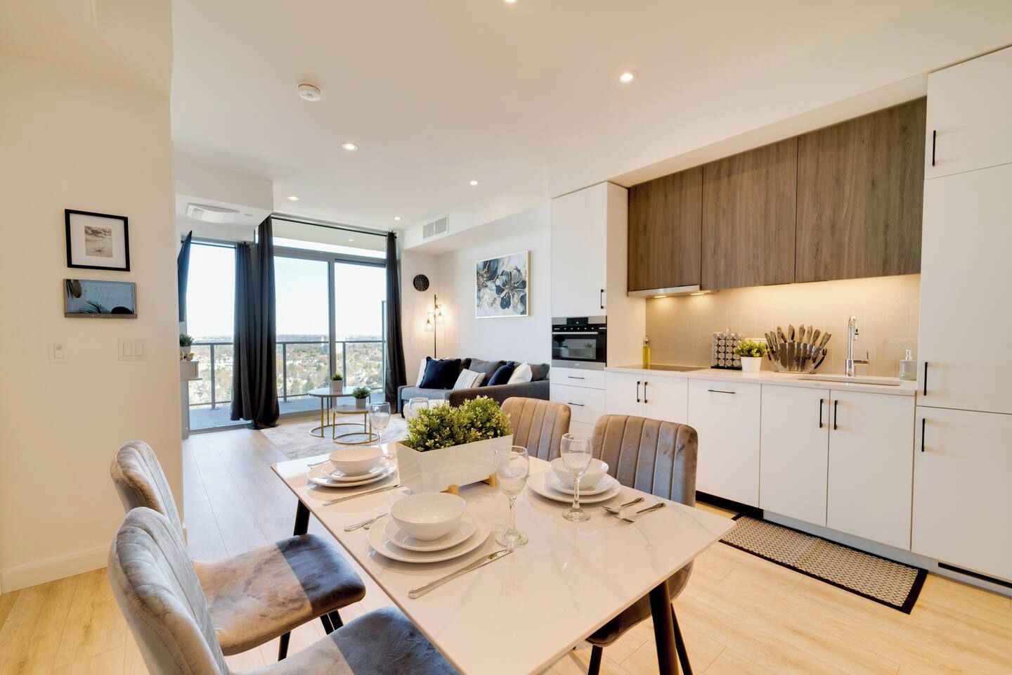 A modern kitchen with white cabinets, a dining area with a table set for four, and a view of the city through large windows.