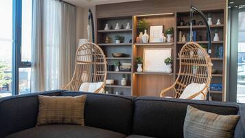 A modern living room with a dark grey sofa, two wicker chairs, a wooden bookshelf, and a large window with curtains.