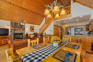 A spacious living area with a fireplace, a dining table with a checkered table runner, and a kitchen with wooden cabinets. - Little Bear Chalet - Mtn Views/Hot Tub/EV Charger (Bryson City)