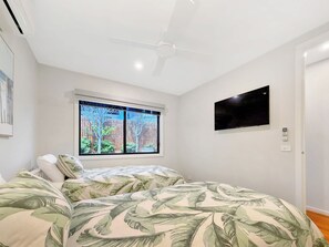A bedroom with a bed, a ceiling fan, a window with a view of trees, and a flat-screen TV mounted on the wall. - Ocean Grove Luxe One Block from Main Beach (Ocean Grove)