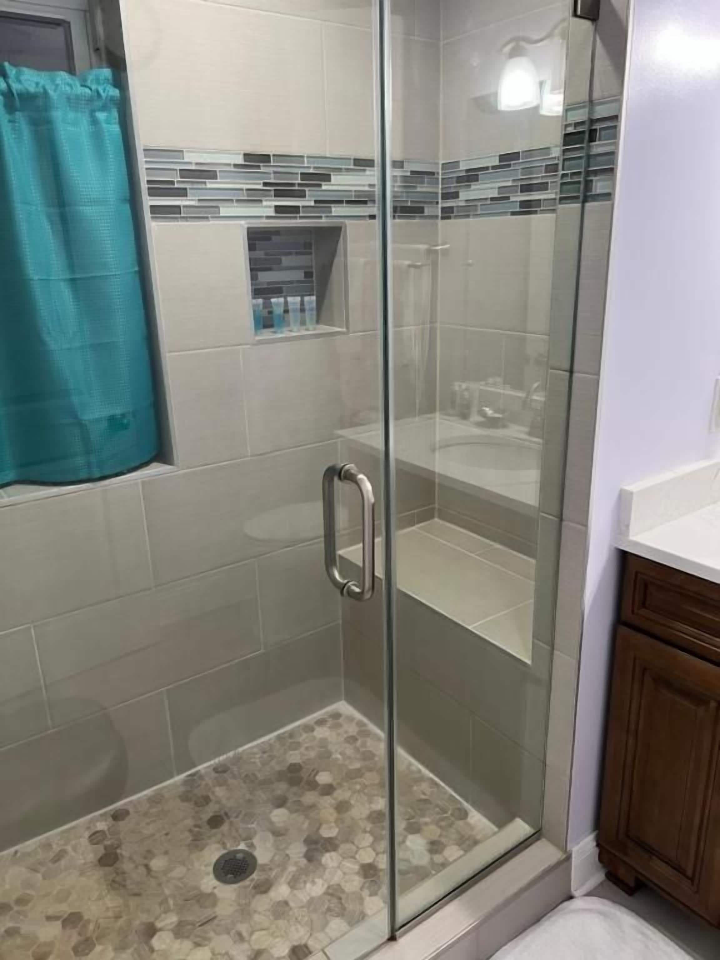A glass-enclosed shower with a tiled floor, a metal handle, and a small shelf with toiletries.