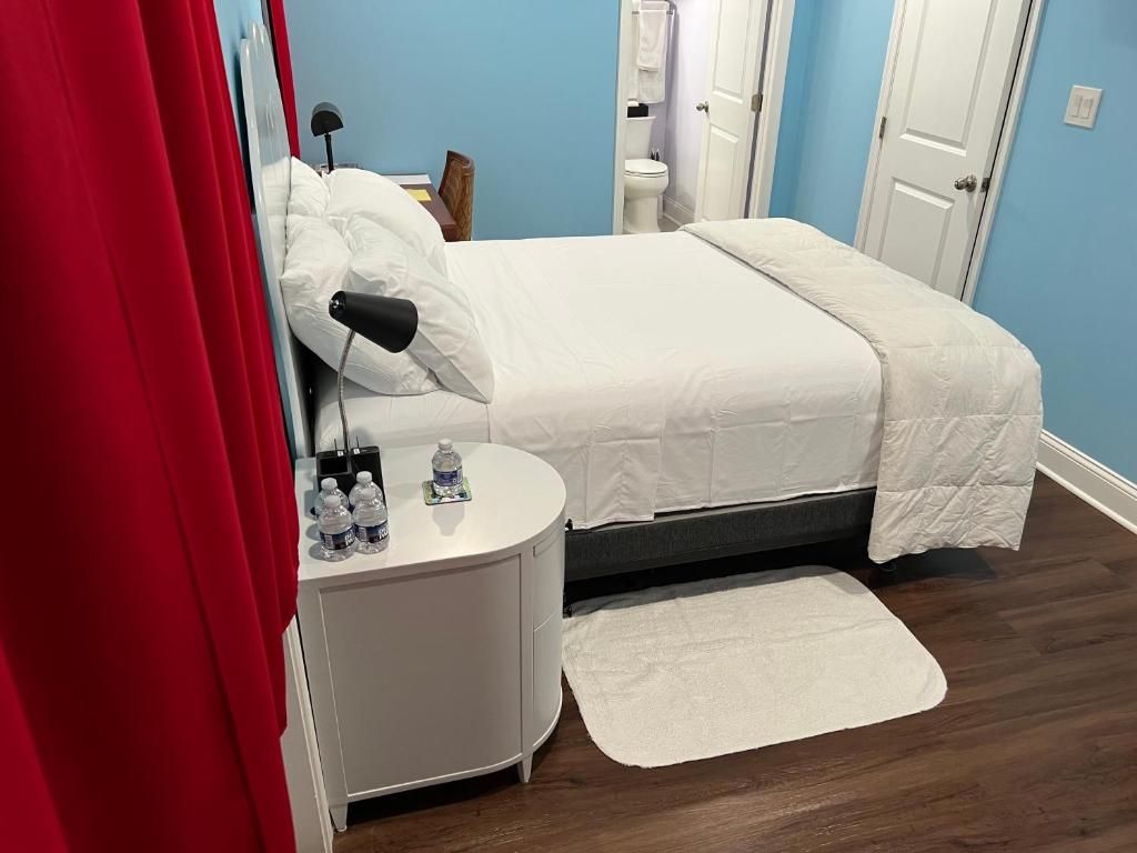Habitación de hotel con cama, mesita de noche con botellas de agua, alfombra blanca y cortina roja.