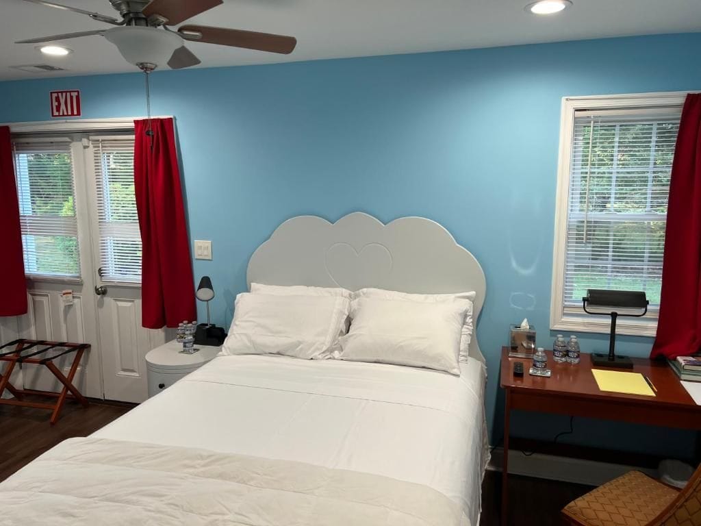 Une chambre à coucher comprenant un lit, un bureau, une chaise, un ventilateur de plafond et une fenêtre avec des stores.