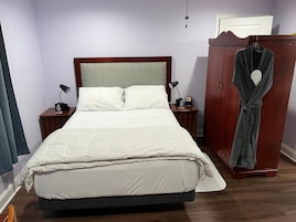 Una cama bien hecha con cabecera de madera, dos mesitas de noche con lámparas, una bata colgada en un armario de madera y una ventana con cortinas.