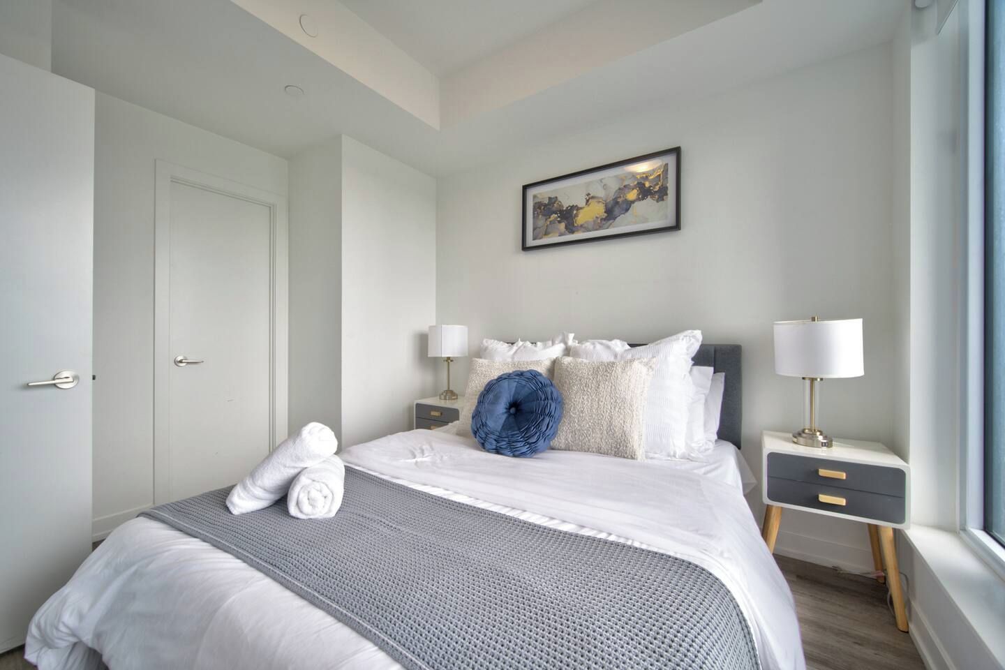 A hotel room with a bed, a bedside table with a lamp, a wall-mounted painting, and a window.