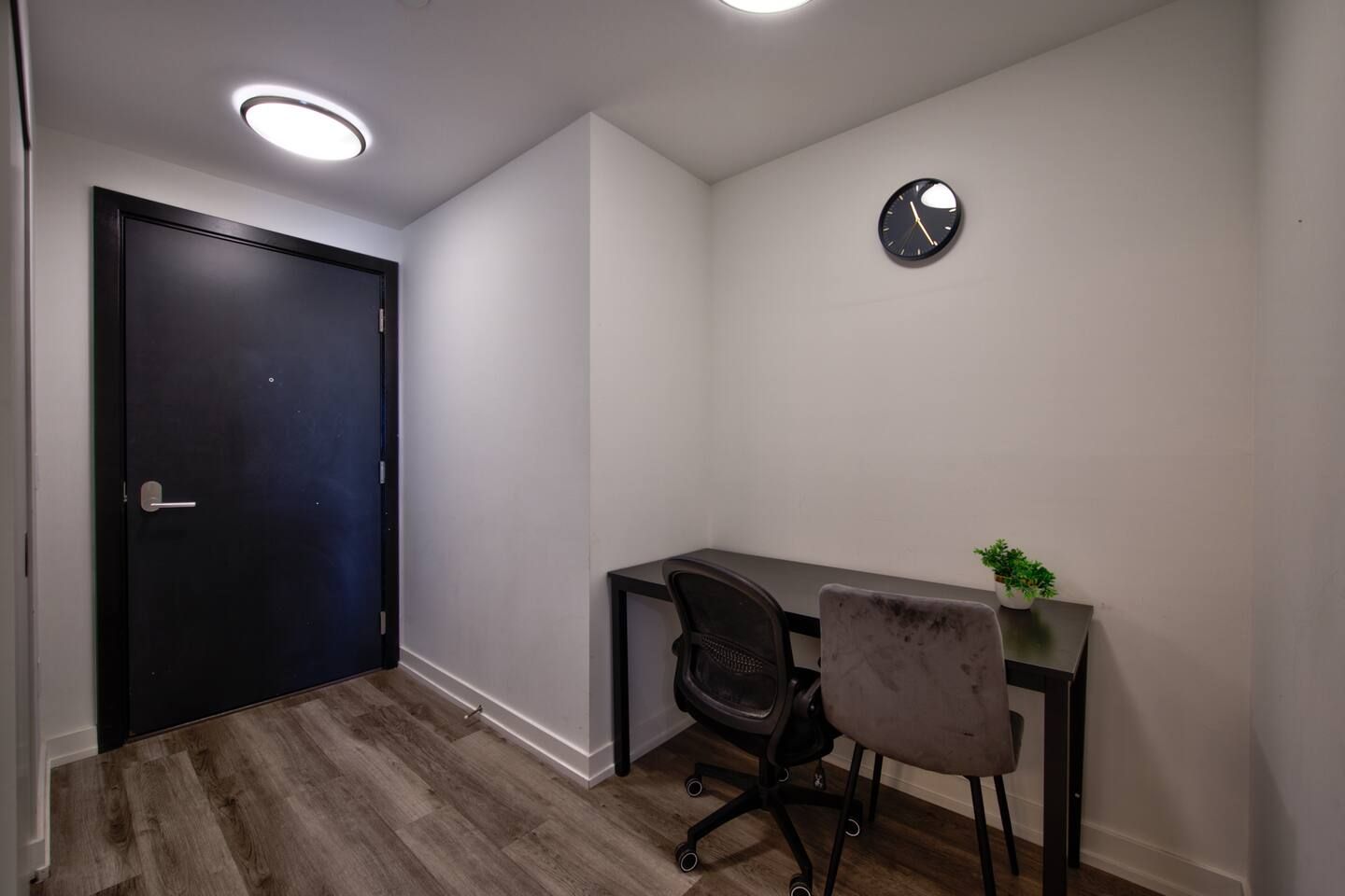 A room with a dark door, a desk with a chair, a small plant, and a wall clock.