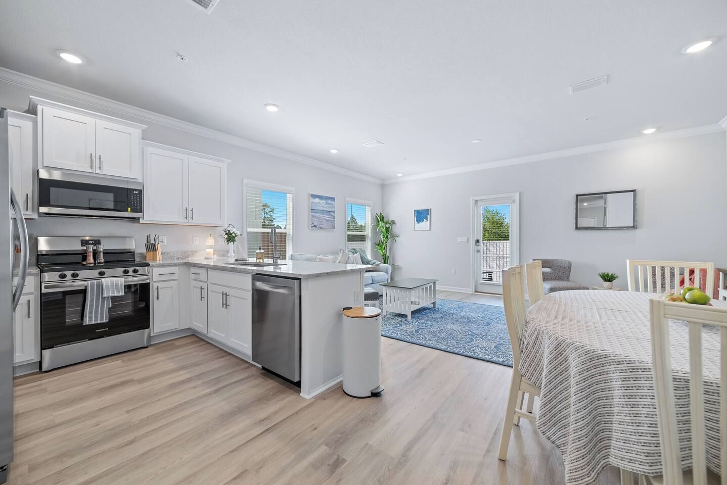 A modern kitchen with white cabinetry, a stainless steel stove, and a dishwasher. There is a dining area with a table and chairs, and a blue rug on the floor.