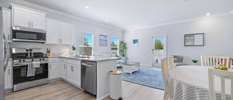 A modern kitchen with white cabinetry, a stainless steel stove, and a dishwasher. There is a dining area with a table and chairs, and a blue rug on the floor.